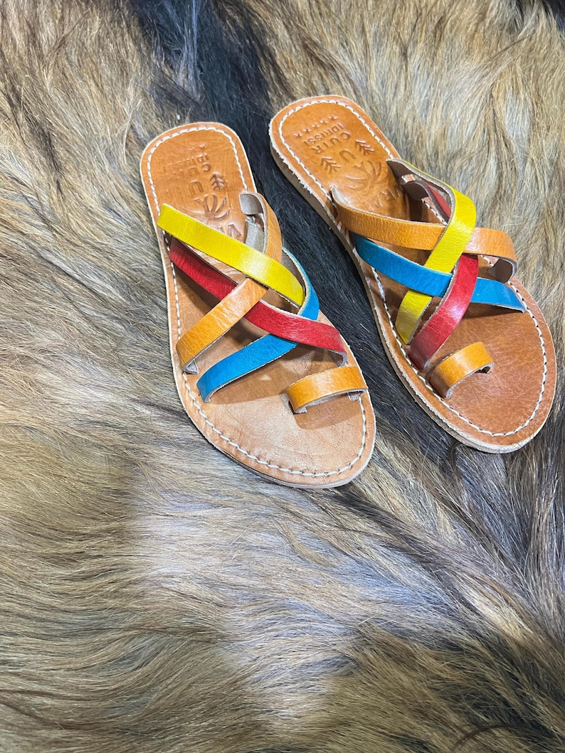 K&ouml;nnte beinhalten: Ein Paar handgefertigte Ledersandalen mit bunten Riemen. Die Sandalen haben eine braune Ledersohle mit Zehenschlaufe und gekreuzten Riemen in Gelb, Rot, Blau und Orange. Die Sandalen sind mit wei&szlig;em Faden gen&auml;ht.