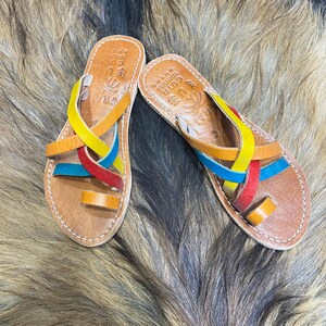 K&ouml;nnte beinhalten: Ein Paar handgefertigte Ledersandalen mit bunten Riemen in Gelb, Orange, Rot und Blau. Die Sandalen haben eine Zehenschlaufe und sind an den Kanten gen&auml;ht. Die Sohlen sind hellbraun. Die Sandalen haben Text auf der Innensohle.