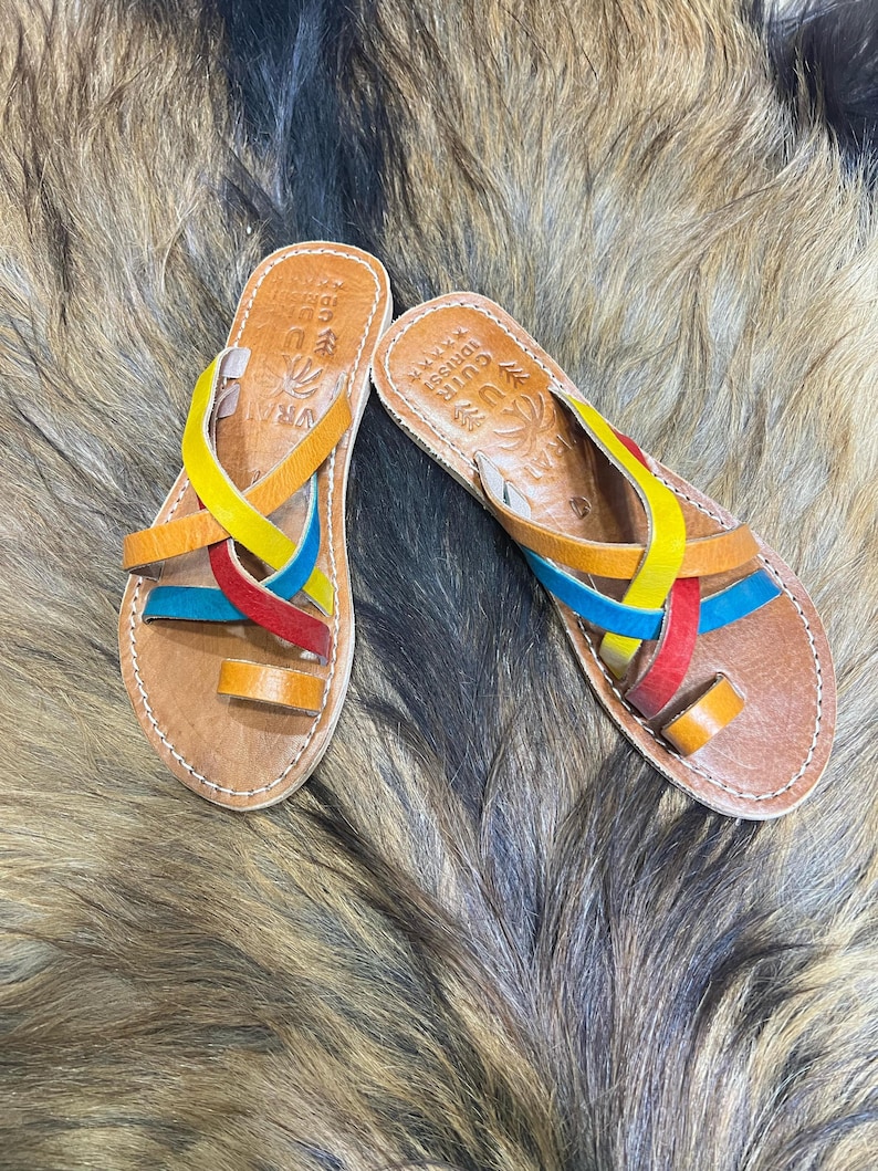K&ouml;nnte beinhalten: Ein Paar handgefertigte Ledersandalen mit bunten Riemen in Gelb, Orange, Rot und Blau. Die Sandalen haben eine Zehenschlaufe und sind an den Kanten gen&auml;ht. Die Sohlen sind hellbraun. Die Sandalen haben Text auf der Innensohle.