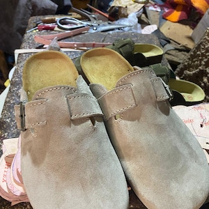 May include: A pair of light grey suede clogs with buckle fastenings. The clogs have a tan interior and are on a work surface with other shoe components and tools in the background. The clogs are a neutral colour and appear to be handmade.