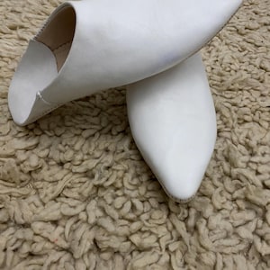 Op de afbeelding: Een paar witte leren slippers met spitse neuzen. De slippers liggen op een getextureerd, beige tapijt. Het schoeisel heeft een eenvoudig, elegant ontwerp, geschikt voor binnenshuis.