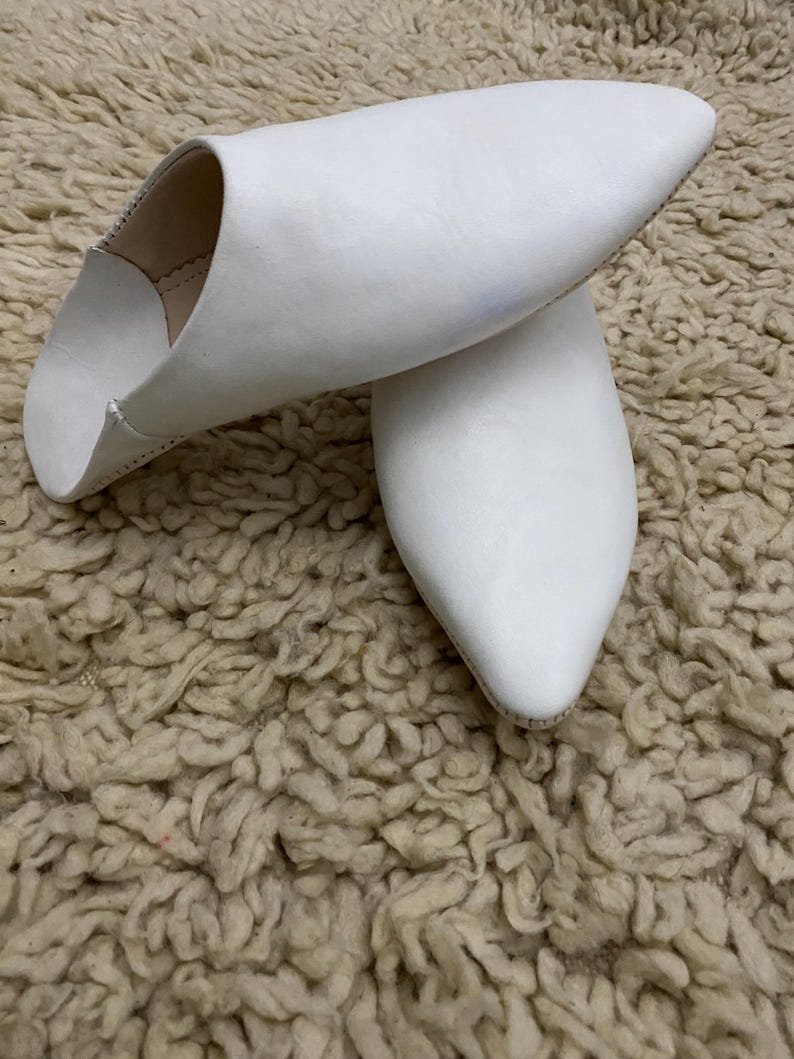 Op de afbeelding: Een paar witte leren slippers met spitse neuzen. De slippers liggen op een getextureerd, beige tapijt. Het schoeisel heeft een eenvoudig, elegant ontwerp, geschikt voor binnenshuis.