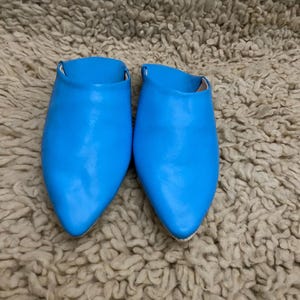 Op de afbeelding: Een paar felblauwe leren slippers met spitse neuzen. De schoenen staan op een getextureerd, lichtbruin tapijt. De slippers hebben een eenvoudig instapontwerp en zijn gemaakt van glad, glanzend materiaal.