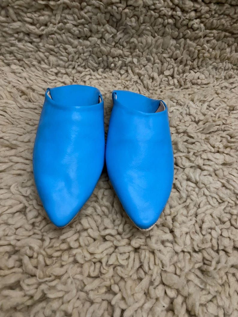 Op de afbeelding: Een paar felblauwe leren slippers met spitse neuzen. De schoenen staan op een getextureerd, lichtbruin tapijt. De slippers hebben een eenvoudig instapontwerp en zijn gemaakt van glad, glanzend materiaal.