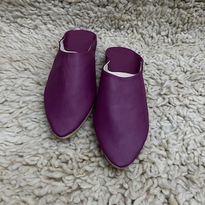 Op de afbeelding: Een paar paarse leren slippers met een puntige neus. De slippers zijn een diepe paarse tint en hebben een eenvoudig, instapontwerp. De binnenkant is wit. De slippers liggen op een getextureerd, gebroken wit tapijt.