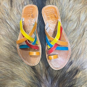 K&ouml;nnte beinhalten: Ein Paar Ledersandalen mit bunten Riemen in Gelb, Rot, Blau und Orange. Die Sandalen haben eine Zehenschlaufe und gekreuzte Riemen. Das Fu&szlig;bett ist hellbraun mit gepr&auml;gtem Text und einem dekorativen Design.
