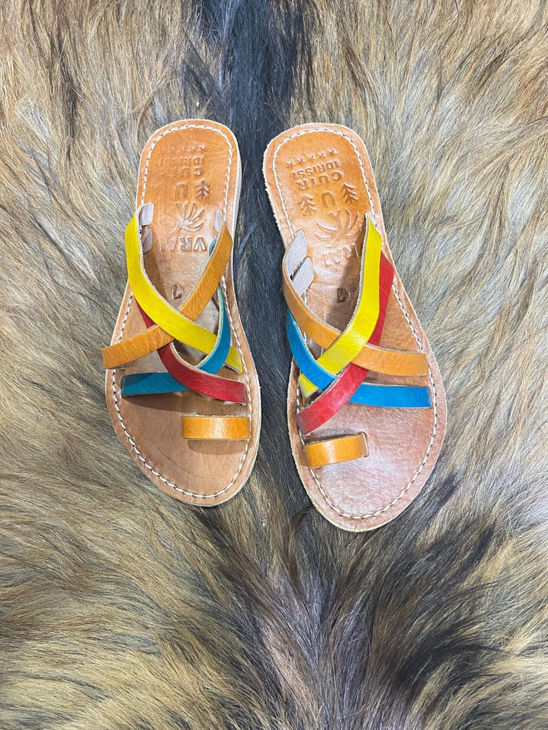 K&ouml;nnte beinhalten: Ein Paar Ledersandalen mit bunten Riemen in Gelb, Rot, Blau und Orange. Die Sandalen haben eine Zehenschlaufe und gekreuzte Riemen. Das Fu&szlig;bett ist hellbraun mit gepr&auml;gtem Text und einem dekorativen Design.