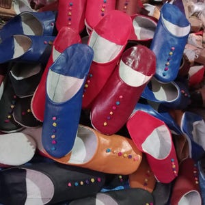 Puede incluir: Una colección de zapatillas de cuero de colores, en varios tonos, incluyendo rojo, azul y negro. Cada zapatilla presenta una fila de pequeños botones decorativos en colores contrastantes. Las zapatillas son de estilo tradicional, probablemente para uso en interiores.