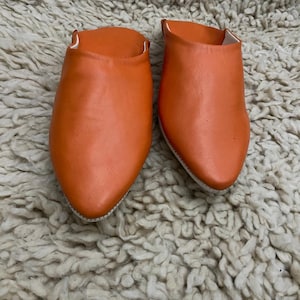 Op de afbeelding: Een paar oranje leren slippers met spitse tenen. De slippers hebben een levendige oranje tint en lijken handgemaakt. De binnenkant is wit. De slippers worden gepresenteerd op een getextureerd, gebroken wit tapijt.