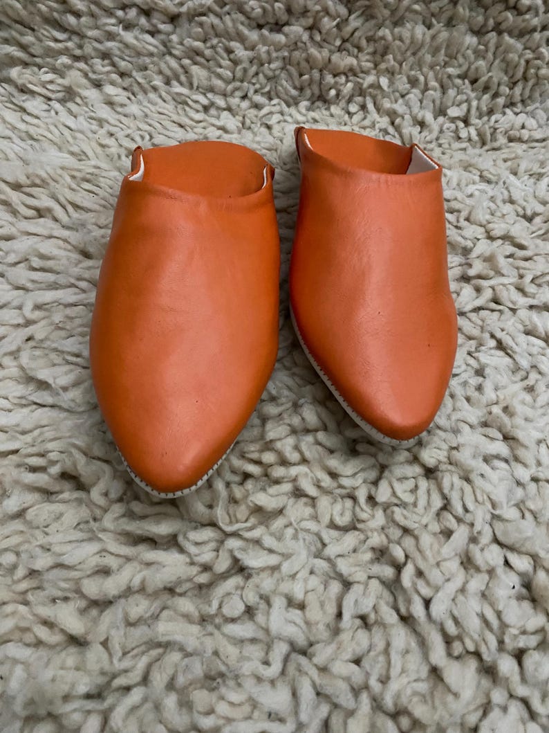Op de afbeelding: Een paar oranje leren slippers met spitse tenen. De slippers hebben een levendige oranje tint en lijken handgemaakt. De binnenkant is wit. De slippers worden gepresenteerd op een getextureerd, gebroken wit tapijt.