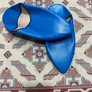 Puede incluir: Un par de zapatillas de cuero azul con punta puntiaguda. Las zapatillas están sobre una alfombra estampada.