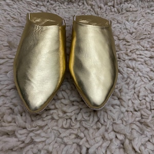 Op de afbeelding: Een paar gouden leren slippers met spitse neuzen. De slippers hebben een metallic glans en liggen op een getextureerde, gebroken witte achtergrond. De schoenen lijken nieuw.