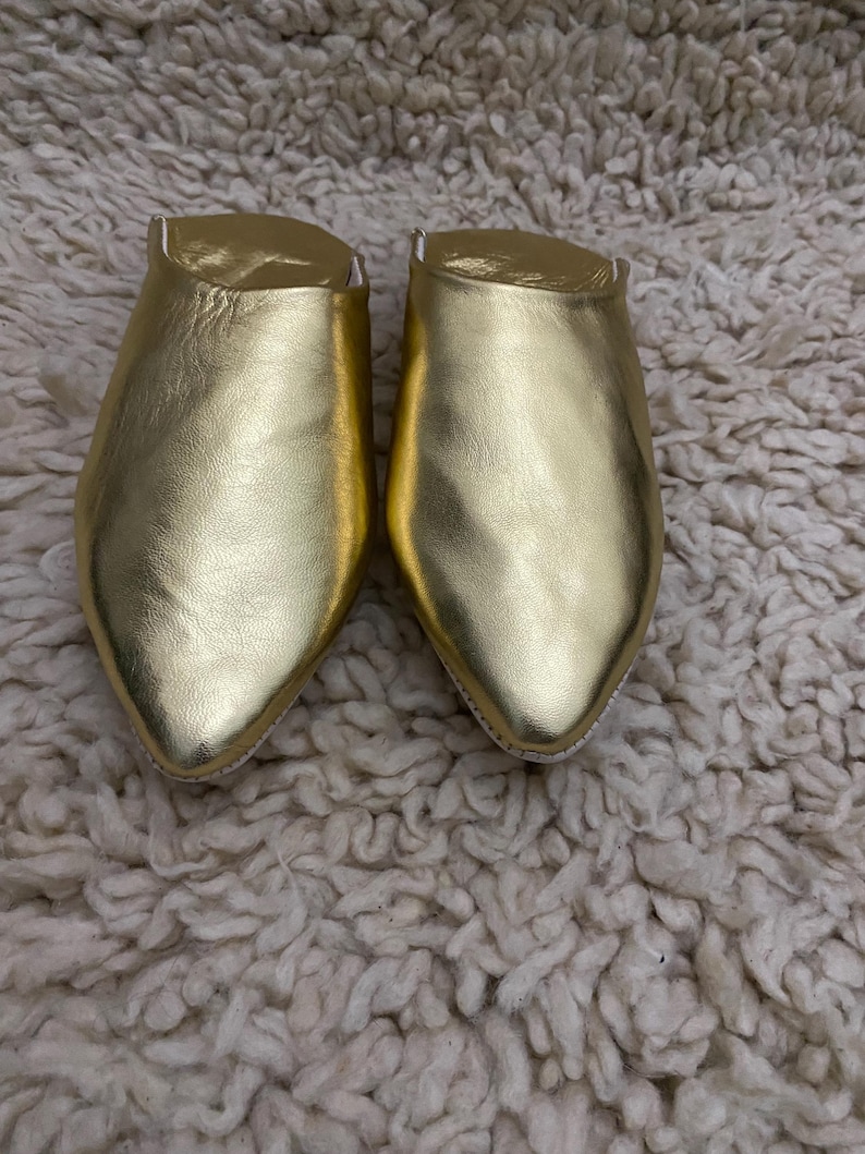 Op de afbeelding: Een paar gouden leren slippers met spitse neuzen. De slippers hebben een metallic glans en liggen op een getextureerde, gebroken witte achtergrond. De schoenen lijken nieuw.