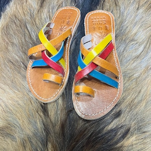 K&ouml;nnte beinhalten: Ein Paar handgefertigte Ledersandalen mit bunten gekreuzten Riemen in Gelb, Rot, Blau und Orange. Die Sandalen haben eine Zehenschlaufe und sind auf einer braunen Ledersohle angebracht. Die Sandalen werden auf einer strukturierten, fellartigen Oberfl&auml;che pr&auml;sentiert.