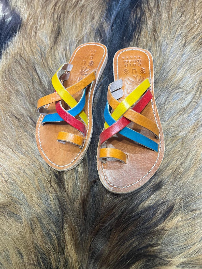 K&ouml;nnte beinhalten: Ein Paar handgefertigte Ledersandalen mit bunten gekreuzten Riemen in Gelb, Rot, Blau und Orange. Die Sandalen haben eine Zehenschlaufe und sind auf einer braunen Ledersohle angebracht. Die Sandalen werden auf einer strukturierten, fellartigen Oberfl&auml;che pr&auml;sentiert.