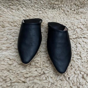 Op de afbeelding: Een paar zwarte leren slippers met spitse neuzen. Het open rugontwerp heeft een gladde, matte afwerking. De slippers worden gepresenteerd op een getextureerd, gebroken wit tapijt.