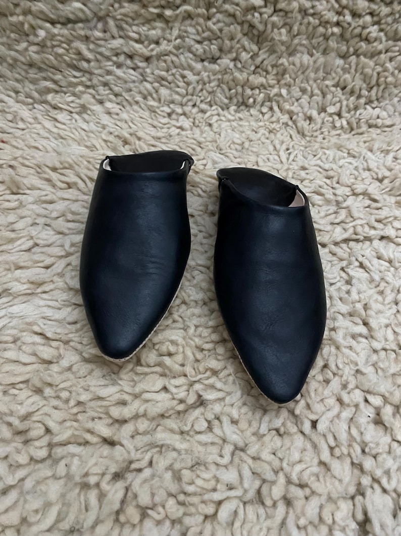 Op de afbeelding: Een paar zwarte leren slippers met spitse neuzen. Het open rugontwerp heeft een gladde, matte afwerking. De slippers worden gepresenteerd op een getextureerd, gebroken wit tapijt.
