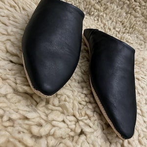 Op de afbeelding: Een paar zwarte leren slippers met een puntige neus en een lichtgekleurde zool. De slippers worden gepresenteerd op een cr&egrave;mekleurig, getextureerd oppervlak, dat hun eenvoudige ontwerp en neutrale kleurenpalet benadrukt.