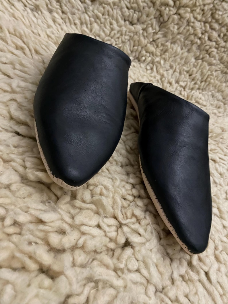 Op de afbeelding: Een paar zwarte leren slippers met een puntige neus en een lichtgekleurde zool. De slippers worden gepresenteerd op een cr&egrave;mekleurig, getextureerd oppervlak, dat hun eenvoudige ontwerp en neutrale kleurenpalet benadrukt.