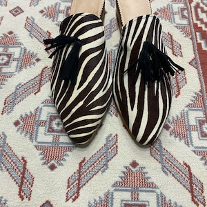 Peut inclure: Une paire de mules en cuir à imprimé zèbre noir et blanc avec des pompons noirs. Les mules sont sur un tapis à motifs.
