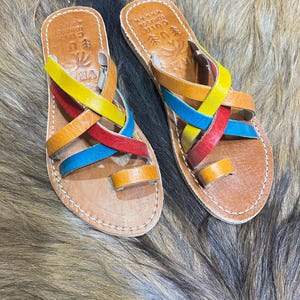 K&ouml;nnte beinhalten: Ein Paar handgefertigte Ledersandalen mit bunten Riemen in Gelb, Rot, Orange und Blau. Die Sandalen haben eine braune Ledersohle mit wei&szlig;en N&auml;hten und eine Zehenschlaufe. Die Sandalen sind mit Text und dekorativen Elementen versehen.