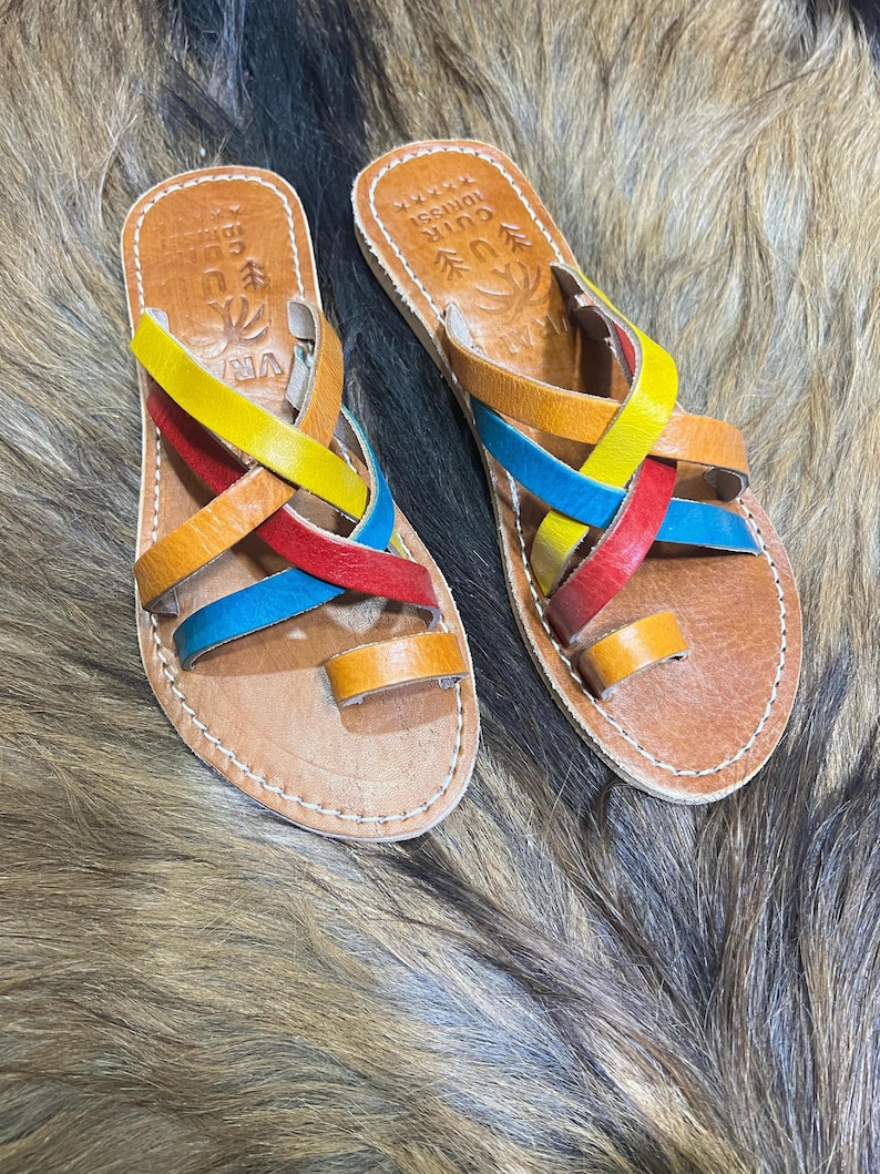 K&ouml;nnte beinhalten: Ein Paar handgefertigte Ledersandalen mit bunten Riemen in Gelb, Rot, Orange und Blau. Die Sandalen haben eine braune Ledersohle mit wei&szlig;en N&auml;hten und eine Zehenschlaufe. Die Sandalen sind mit Text und dekorativen Elementen versehen.