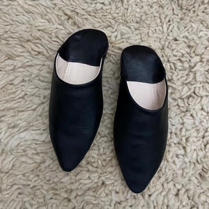 Op de afbeelding: Een paar zwarte leren slippers met spitse neuzen. De binnenkant van de slippers is lichtbeige. De slippers liggen op een getextureerd, gebroken wit tapijt.