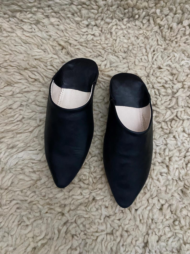 Op de afbeelding: Een paar zwarte leren slippers met spitse neuzen. De binnenkant van de slippers is lichtbeige. De slippers liggen op een getextureerd, gebroken wit tapijt.