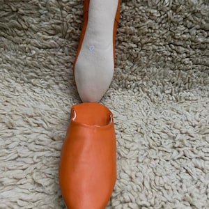 Op de afbeelding: Oranje leren slipper met een puntige neus, getoond met de off-white zool. De slipper wordt getoond op een getextureerd, lichtgekleurd tapijt. De zool is off-white en de bovenkant is effen oranje.