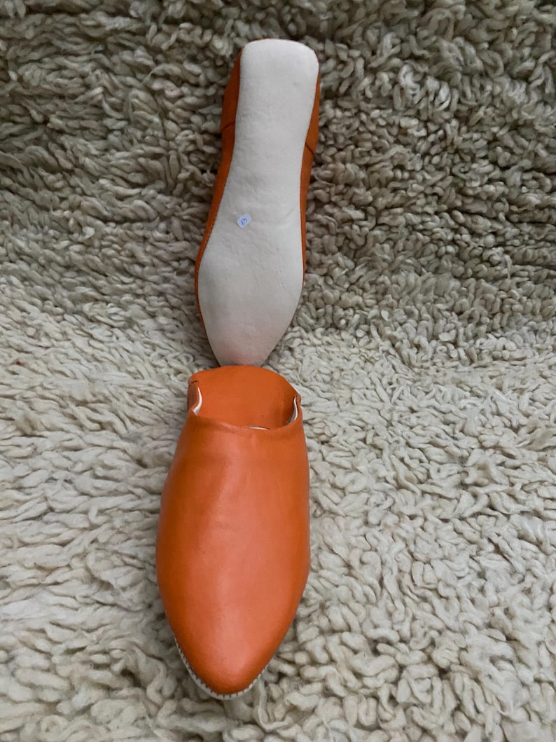 Op de afbeelding: Oranje leren slipper met een puntige neus, getoond met de off-white zool. De slipper wordt getoond op een getextureerd, lichtgekleurd tapijt. De zool is off-white en de bovenkant is effen oranje.