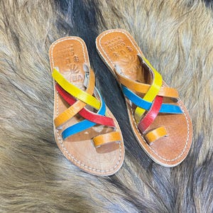 K&ouml;nnte beinhalten: Leder-Sandalen mit bunten Riemen in Gelb, Rot, Blau und Orange. Die Sandalen haben eine Zehenschlaufe und liegen auf einer braunen Oberfl&auml;che.