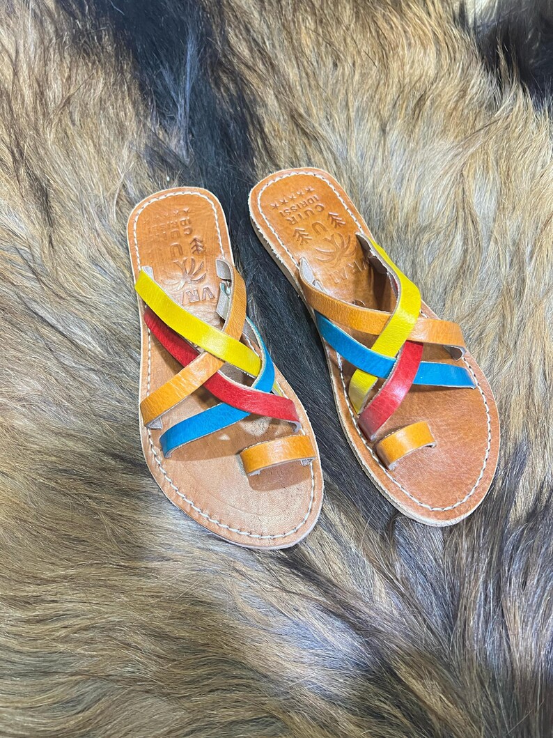 K&ouml;nnte beinhalten: Leder-Sandalen mit bunten Riemen in Gelb, Rot, Blau und Orange. Die Sandalen haben eine Zehenschlaufe und liegen auf einer braunen Oberfl&auml;che.