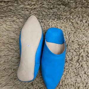 Op de afbeelding: Een paar felblauwe leren slippers met spitse neuzen. E&eacute;n slipper wordt van de zool getoond, met een lichtgekleurde binnenzool en maatmarkering. De andere slipper staat rechtop en toont de levendige blauwe kleur.