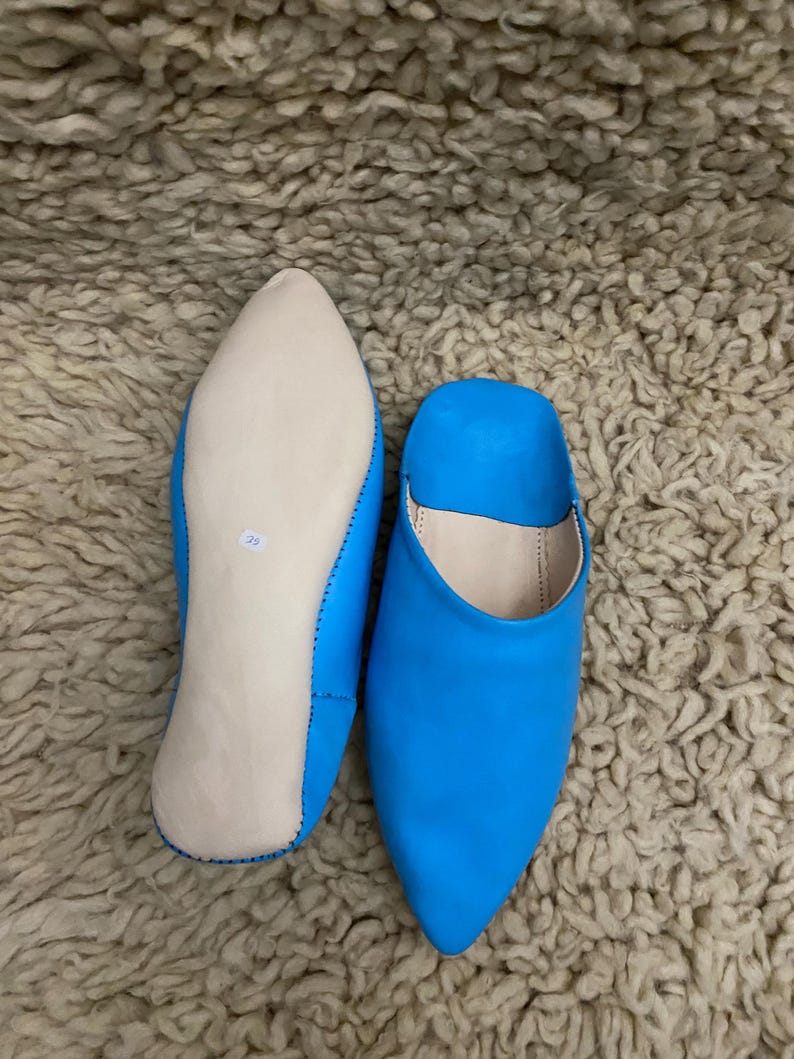 Op de afbeelding: Een paar felblauwe leren slippers met spitse neuzen. E&eacute;n slipper wordt van de zool getoond, met een lichtgekleurde binnenzool en maatmarkering. De andere slipper staat rechtop en toont de levendige blauwe kleur.