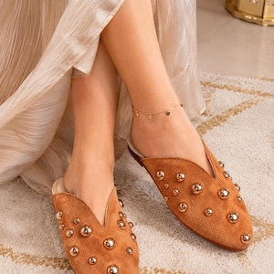 Puede incluir: Un par de zapatos tipo mule de ante color canela con punta afilada y una abertura en forma de V profunda en el empeine. El exterior de cada zapato está decorado con múltiples tachuelas esféricas doradas pulidas sobre el ante.