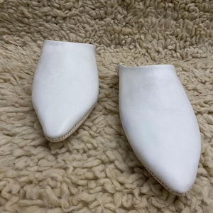 Op de afbeelding: Een paar witte leren slippers met een puntige neus. De slippers zijn gemaakt van glad, effen wit leer en hebben een eenvoudig ontwerp. De zolen zijn lichtbeige van kleur. De slippers zijn waarschijnlijk voor gebruik binnenshuis.
