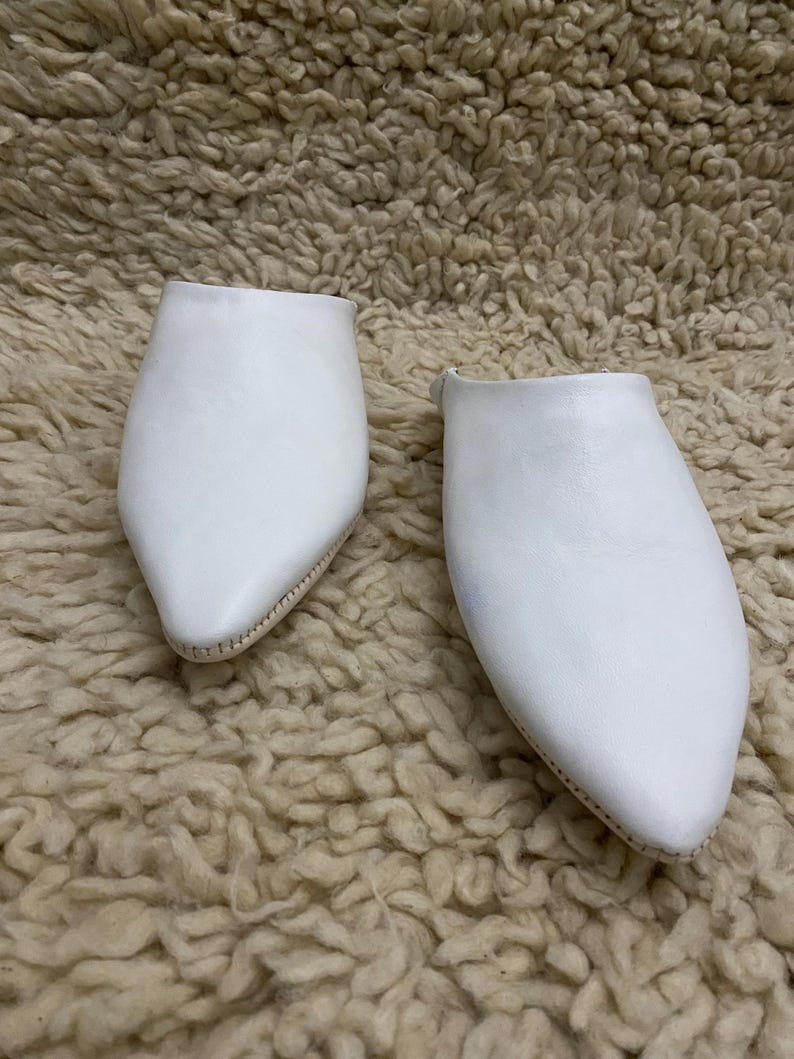 Op de afbeelding: Een paar witte leren slippers met een puntige neus. De slippers zijn gemaakt van glad, effen wit leer en hebben een eenvoudig ontwerp. De zolen zijn lichtbeige van kleur. De slippers zijn waarschijnlijk voor gebruik binnenshuis.