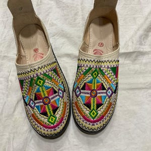 Puede incluir: Par de zapatillas bordadas de color crema. Las zapatillas presentan un colorido diseño geométrico en tonos rosa, verde, azul, amarillo y naranja. El diseño está cosido en la parte delantera de la zapatilla. Las suelas son negras.