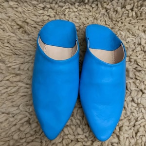 Op de afbeelding: Een paar felblauwe leren slippers met spitse neuzen. De open schoenen hebben een gladde, glanzende afwerking en een neutraal gekleurd interieur. De slippers liggen op een getextureerd, lichtbruin tapijt.