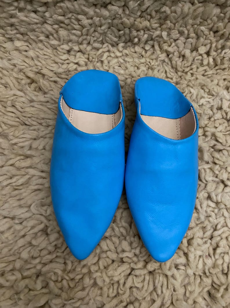 Op de afbeelding: Een paar felblauwe leren slippers met spitse neuzen. De open schoenen hebben een gladde, glanzende afwerking en een neutraal gekleurd interieur. De slippers liggen op een getextureerd, lichtbruin tapijt.
