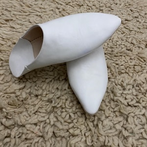 Op de afbeelding: Een paar witte leren slippers met een puntige neus. De slippers zijn gemaakt van glad, wit leer en hebben een eenvoudig, elegant ontwerp. De slippers worden getoond op een getextureerd, beige oppervlak.