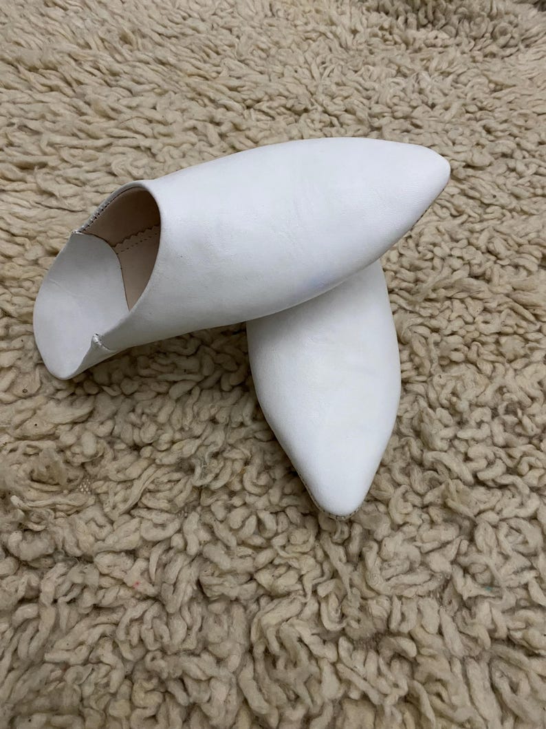 Op de afbeelding: Een paar witte leren slippers met een puntige neus. De slippers zijn gemaakt van glad, wit leer en hebben een eenvoudig, elegant ontwerp. De slippers worden getoond op een getextureerd, beige oppervlak.