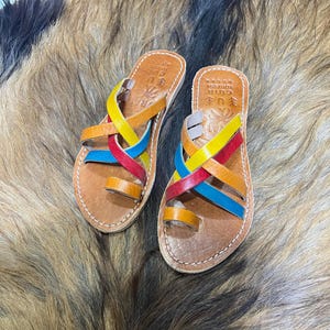 K&ouml;nnte beinhalten: Leder-Sandalen mit farbigen Riemen in Rot, Gelb, Blau und Orange. Die Sandalen haben eine Zehenschlaufe und sind mit wei&szlig;em Faden gen&auml;ht. Die Sohlen sind braun. Der Hintergrund ist eine strukturierte, mehrfarbige Oberfl&auml;che.