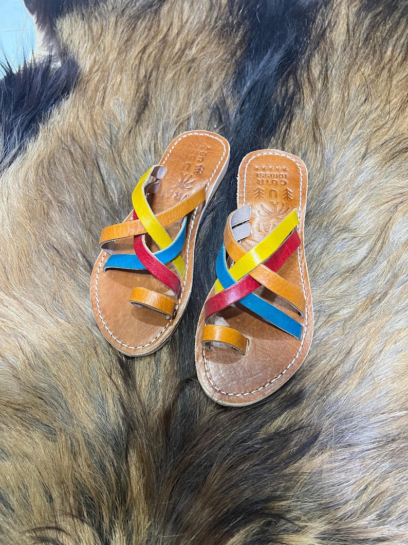 K&ouml;nnte beinhalten: Leder-Sandalen mit farbigen Riemen in Rot, Gelb, Blau und Orange. Die Sandalen haben eine Zehenschlaufe und sind mit wei&szlig;em Faden gen&auml;ht. Die Sohlen sind braun. Der Hintergrund ist eine strukturierte, mehrfarbige Oberfl&auml;che.