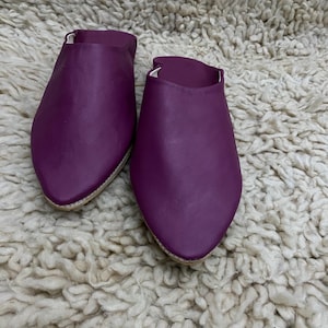 Op de afbeelding: Een paar paarse leren slippers met een spitse neus. De slippers worden gepresenteerd op een getextureerd, gebroken wit tapijt. De slippers hebben een eenvoudig instapontwerp en het leer ziet er glad uit.