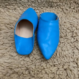 Op de afbeelding: Een paar felblauwe leren slippers met spitse tenen. De slippers staan op een getextureerd, beige tapijt. Het open ontwerp en de levendige kleur maken deze slippers tot een stijlvolle schoenoptie.
