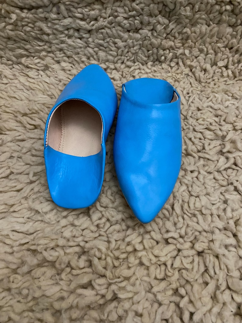 Op de afbeelding: Een paar felblauwe leren slippers met spitse tenen. De slippers staan op een getextureerd, beige tapijt. Het open ontwerp en de levendige kleur maken deze slippers tot een stijlvolle schoenoptie.