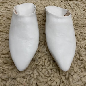 Op de afbeelding: Een paar witte leren instappers met spitse neuzen. De schoenen staan op een getextureerd, lichtbruin tapijt. Het schoeisel heeft een eenvoudig, minimalistisch ontwerp, geschikt voor vrijetijdskleding.