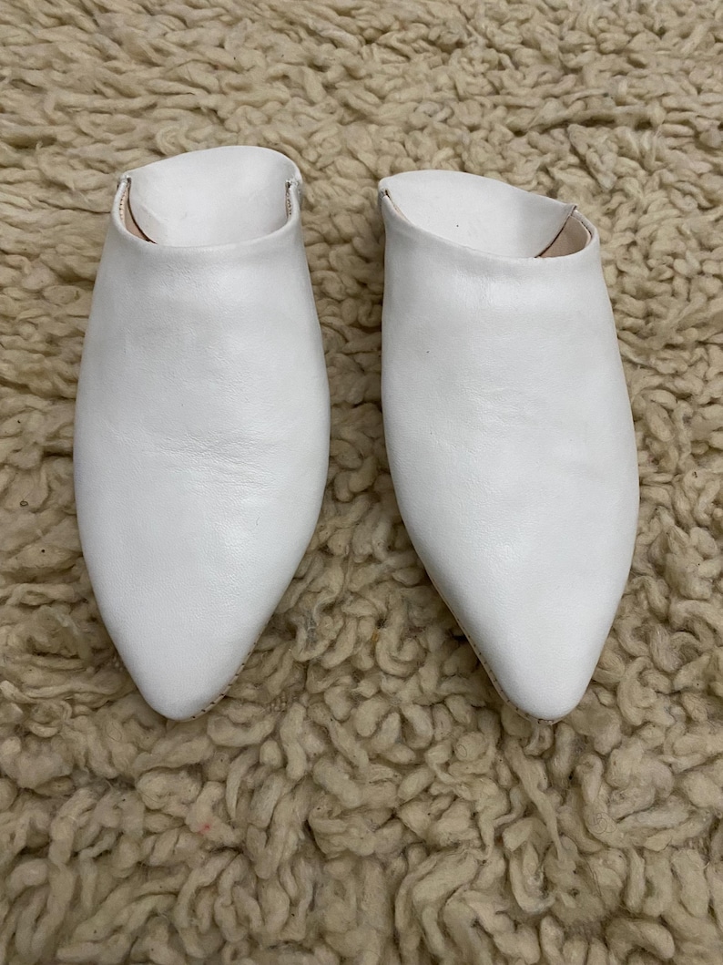 Op de afbeelding: Een paar witte leren instappers met spitse neuzen. De schoenen staan op een getextureerd, lichtbruin tapijt. Het schoeisel heeft een eenvoudig, minimalistisch ontwerp, geschikt voor vrijetijdskleding.