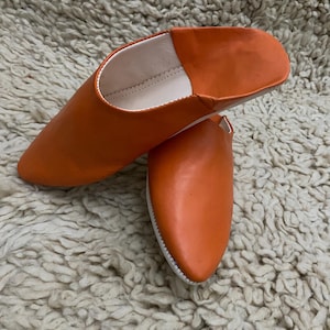 Op de afbeelding: Een paar oranje leren slippers met een puntige neus. De slippers hebben een witte binnenkant en zool. Het schoeisel wordt gepresenteerd op een getextureerd, gebroken wit tapijt.